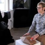 Adult male standing with hands on a customized joystick and trackball controller and speakers and video displays in the background.